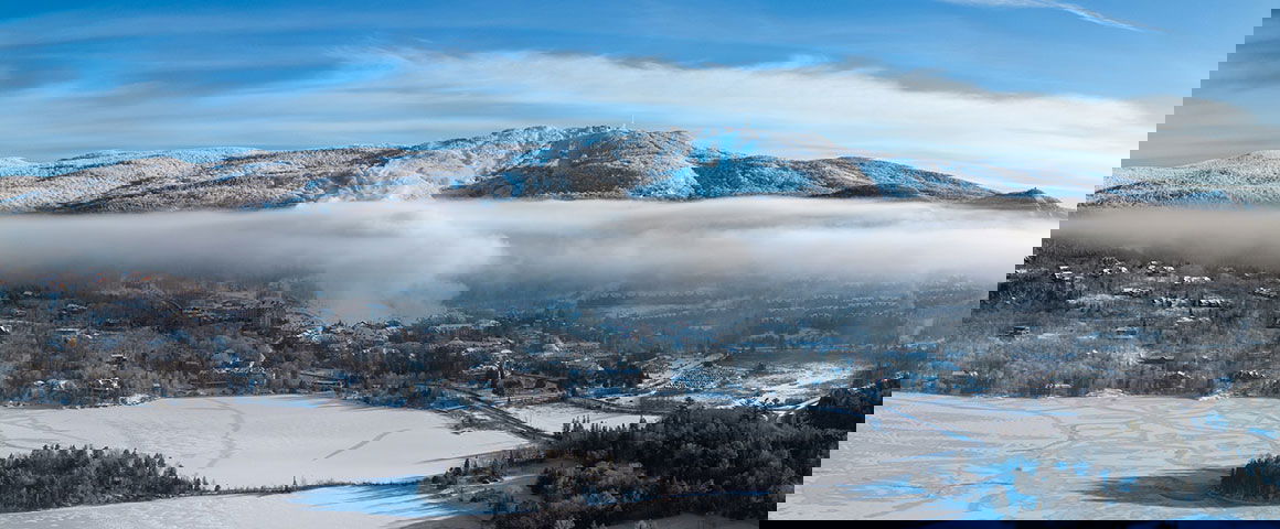 Vue panoramique dun paysage enneigé avec une montagne en arrière-plan. Une couche de brouillard flotte au-dessus dune petite ville nichée dans la vallée. Au premier plan se trouvent des étendues deau gelées entourées darbres recouverts de neige. Un ciel bleu clair au-dessus.